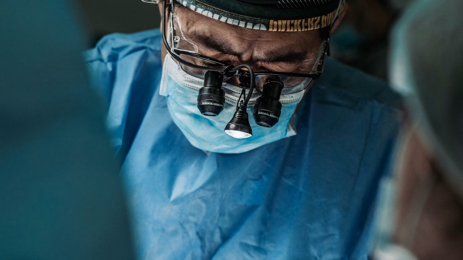 A surgeon in an operating theatre, head bowed in concentration, wearing scrubs, a mask and magnifying loupes with a headlamp