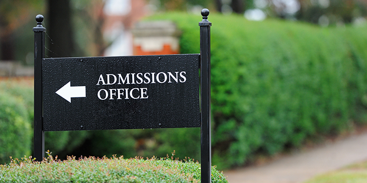 A black "Admissions Office" sign with a white directional arrow, set against green hedges on a university campus.