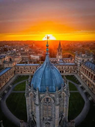 An aerial view over Oxford at sunset, with a college's domed tower in the foreground and the city's spires on the horizon.