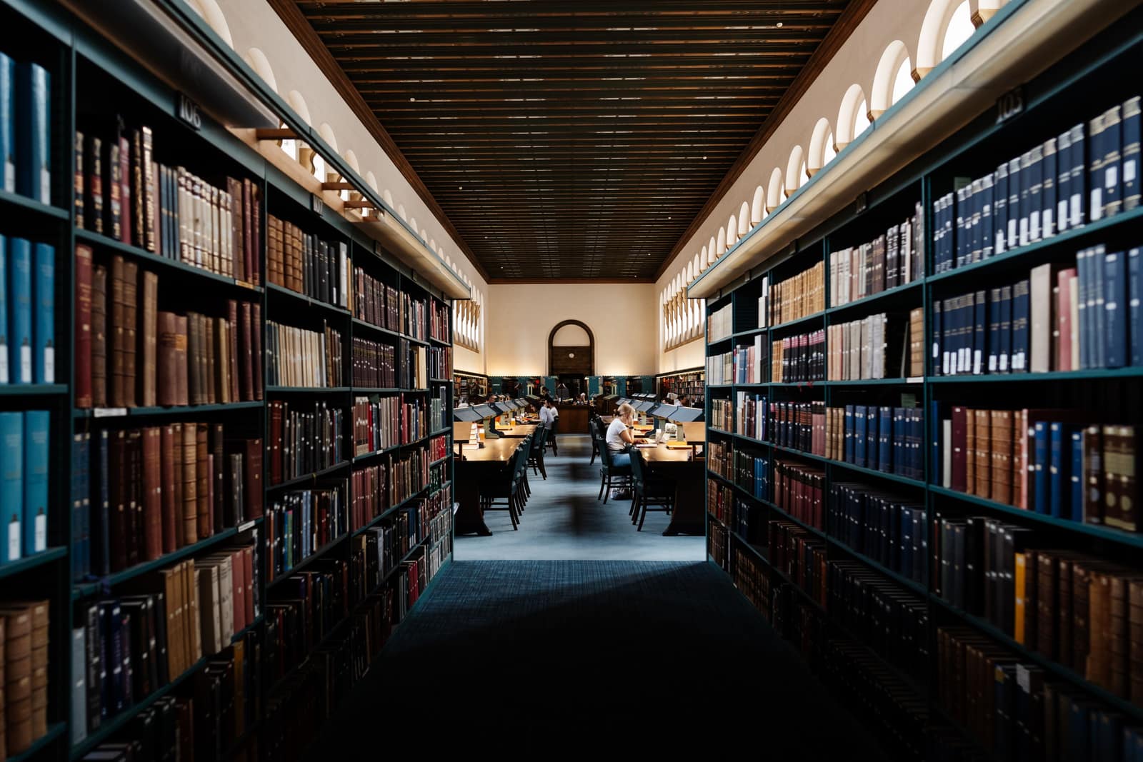 A long library aisle lined with tall shelves of leather-bound books, with readers seated at desks in the distance.