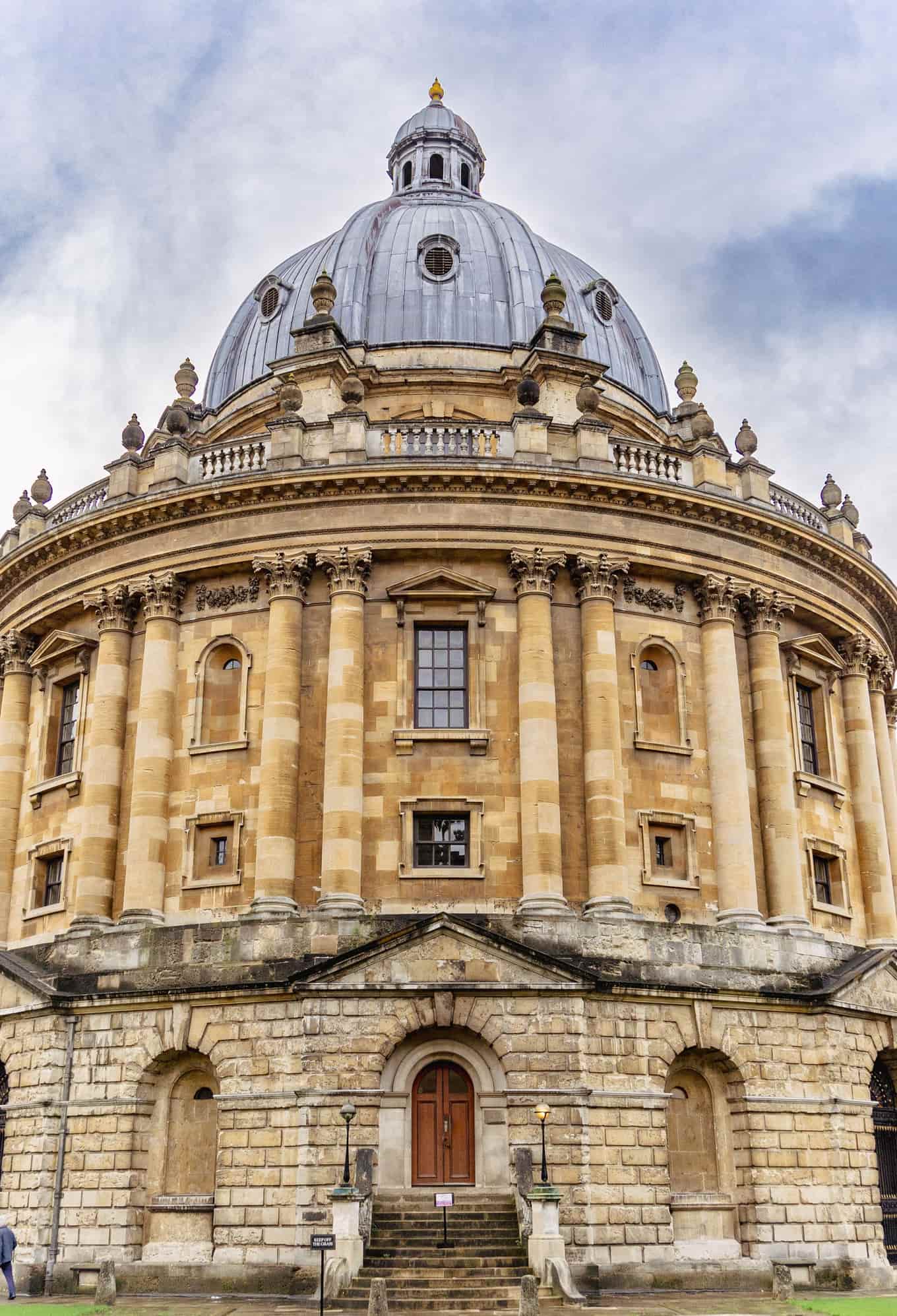 The honey-coloured stone rotunda and lead dome of Oxford's Radcliffe Camera seen from below against a cloudy sky.