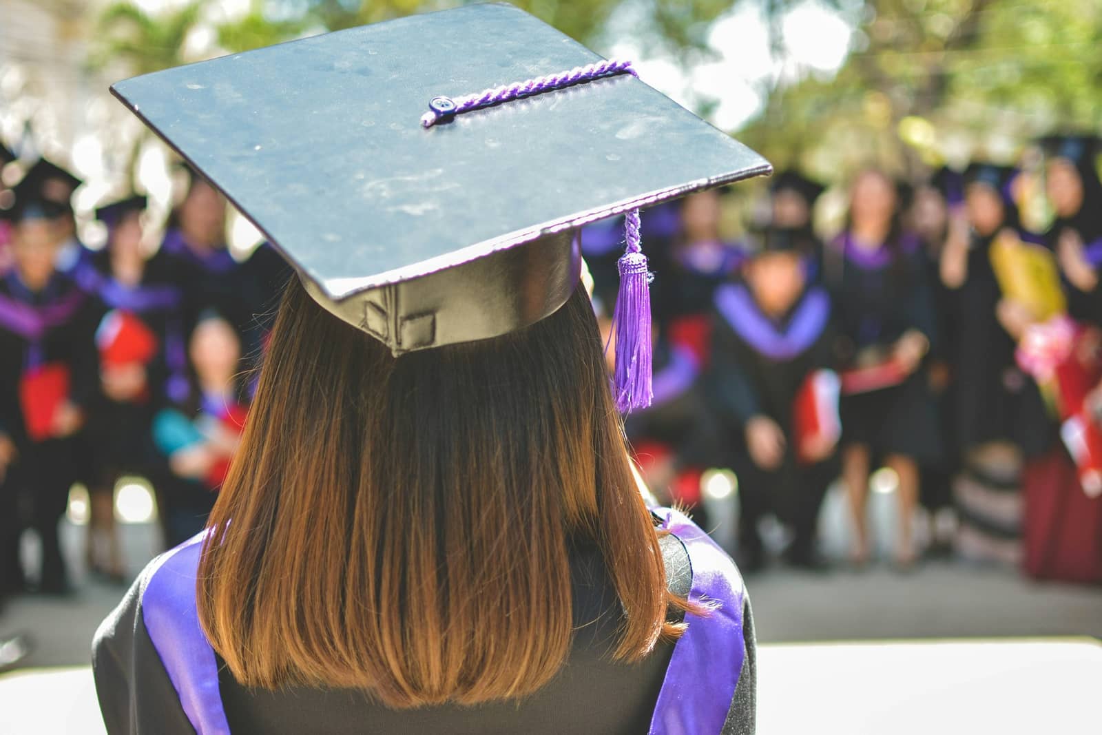 A graduate seen from behind wearing a mortarboard and gown, facing a blurred crowd of fellow graduates at a commencement ceremony.