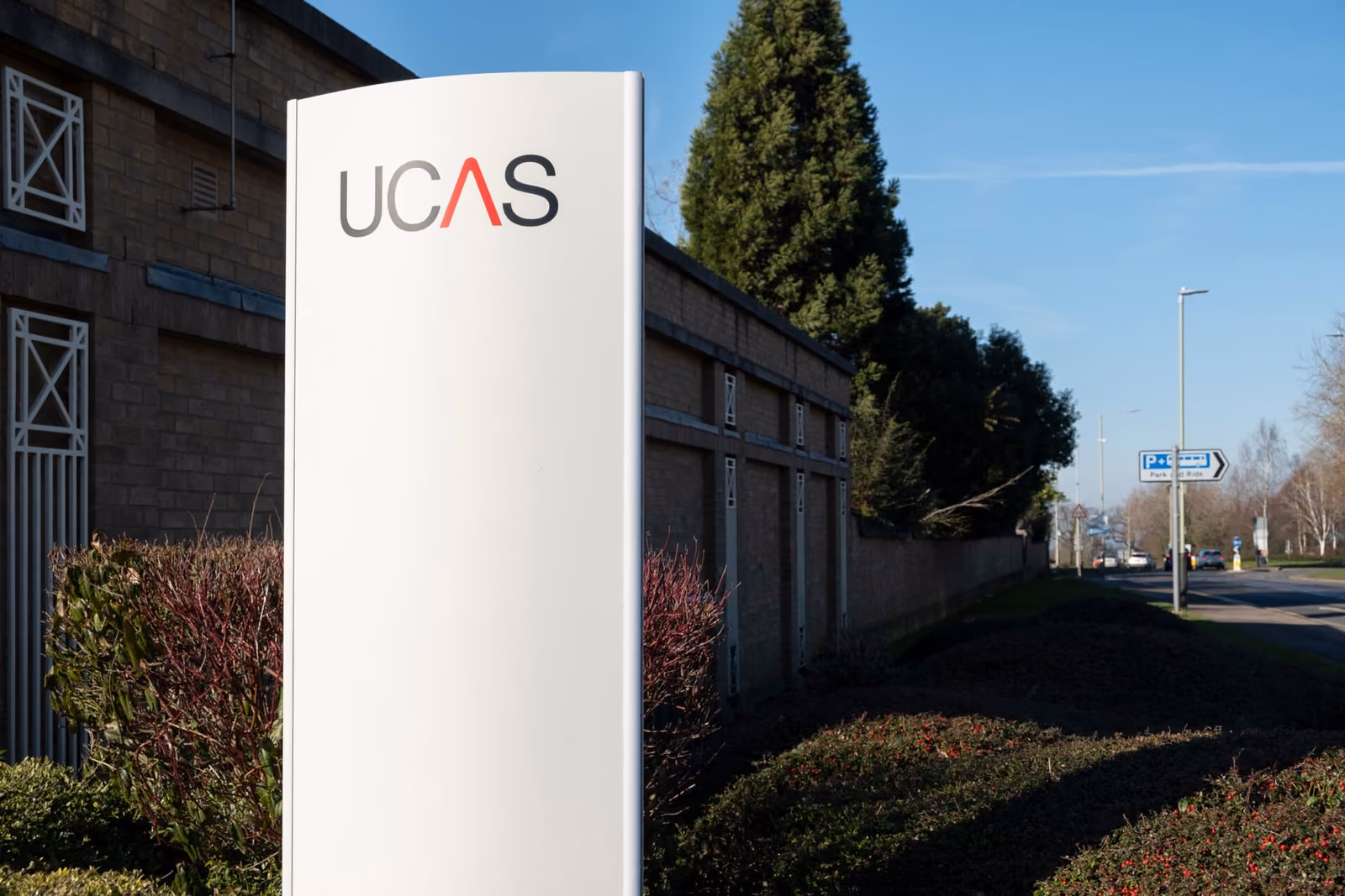 A white UCAS sign standing beside a road outside a brick building.